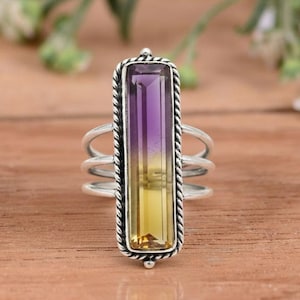 May include: A silver ring with a rectangular, two-tone gemstone in shades of purple and yellow. The stone is set in a textured bezel, and the ring has a triple-band design.