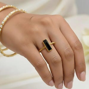 Könnte beinhalten: Ein goldfarbener Ring mit einem rechteckigen schwarzen Stein wird auf einer Hand gezeigt. Zwei goldene Armreifen mit kleinen, klaren Steinen sind ebenfalls zu sehen. Der Schmuck ist vor einem weichen, neutralen Hintergrund platziert.