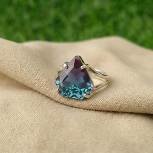 Può includere: Un anello d'argento con una grande gemma sfaccettata con una sfumatura di colore dal viola scuro al turchese. La gemma è incastonata in una fascia a gambo diviso, su uno sfondo di tessuto beige. Il design dell'anello è elegante e accattivante.