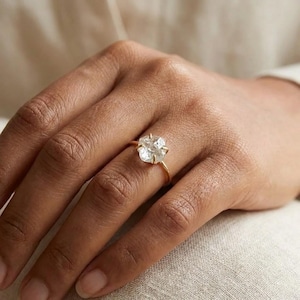 Könnte beinhalten: Ein goldener Ring mit einem großen, unregelmäßig geformten, klaren Edelstein. Der Ring hat ein dünnes, gelbes Band, und der Edelstein ist in einer Krappenfassung gehalten. Der Ring wird an einem Finger getragen.