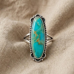 May include: A silver ring with a large, oval turquoise stone. The stone has brown veining and is set in a decorative bezel with a beaded edge. The ring has a split shank and a vintage style.