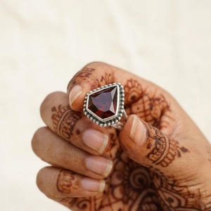 Peut inclure: Une bague en argent ornée d'une grosse pierre précieuse rouge foncé facettée. La pierre est sertie dans une forme géométrique avec une bordure perlée. La bague est tenue dans une main avec des motifs au henné.