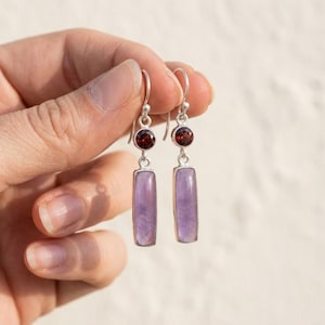 May include: A pair of silver drop earrings featuring a small, round, deep red garnet gemstone set above a longer, rectangular, light purple amethyst stone. The earrings hang from simple silver hooks and have a polished metal finish.