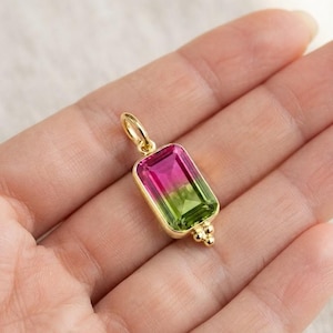 Puede incluir: Un colgante rectangular con una piedra preciosa de turmalina sandía facetada con un degradado de tonos rosa y verde vibrantes, engastada en un marco de metal dorado con una anilla y tres pequeñas cuentas doradas en la base.