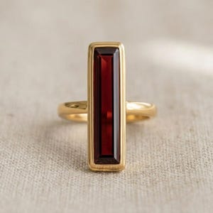 May include: A gold ring featuring a rectangular, deep red gemstone. The gemstone is set vertically within a gold bezel. The ring's band is a simple, polished gold.