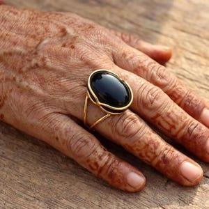 May include: A gold-colored ring featuring a large, oval-shaped black stone. The ring's design includes a double-banded setting that cradles the stone. The ring is displayed on a hand, showcasing the jewelry's design and color.