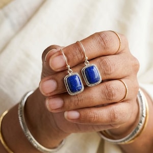May include: A pair of drop earrings featuring rectangular lapis lazuli stones set in silver frames with a twisted rope border. The stones are a deep blue color with subtle natural inclusions, suspended from simple silver ear wires.