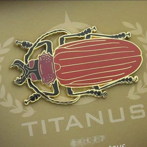 May include: A red and gold beetle pin with a detailed design. The pin features a red body with gold stripes and a gold outline. The beetle is set against a tan background with the word "TITANUS" in white.