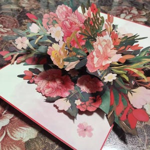 May include: A pop-up greeting card featuring a vibrant floral arrangement. The card showcases detailed paper cutouts of pink peonies, red leaves, and various other flowers. The card is open, revealing the three-dimensional bouquet.