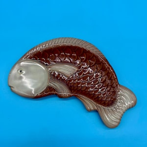 May include: A ceramic fish-shaped dish. The body is brown, and the head and fins are light grey. The fish has a textured scale pattern. It is set against a bright blue background. The dish is likely for decorative or serving purposes.