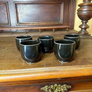 Könnte beinhalten: Fünf schwarze Keramikbecher mit glänzendem Finish sind auf einer Holzoberfläche angeordnet. Die Becher haben eine abgerundete Form mit einem leicht ausgestellten Rand. Der Hintergrund zeigt einen dunklen Holzschrank mit dekorativen Elementen.