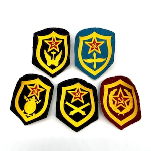 May include: A collection of six military patches. Each patch features a shield shape with a yellow border and a red star emblem. The patches have various designs, including wings, cannons, and a wreath, set against backgrounds of black, blue, and maroon.
