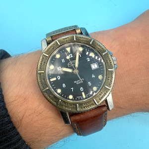 Puede incluir: Un reloj de pulsera con esfera negra, manecillas luminosas y ventana de fecha. El reloj tiene un bisel color bronce con nombres de ciudades del mundo y una correa de cuero marrón. La esfera del reloj dice "QUARTZ 10ATM".