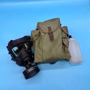 Puede incluir: Una máscara de gas negra con lentes transparentes, una bolsa de lona verde y una botella de agua de plástico blanco se muestran sobre un fondo azul. La máscara de gas tiene un filtro adjunto. La bolsa tiene un cierre de solapa.