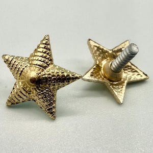 May include: Two gold-colored star-shaped cabinet knobs. The knobs have a textured surface and a screw post on the back. The star points are slightly rounded. These decorative knobs can be used for furniture or other DIY projects.