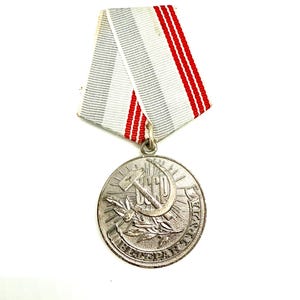May include: A silver-colored medal with a hammer and sickle emblem, laurel branches, and Cyrillic text. The medal hangs from a striped ribbon with gray, white, and red stripes, against a white background.
