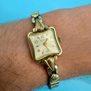 May include: A vintage gold-coloured wristwatch with a square face and a gold band. The watch face displays the numbers 10, 12, 2, 4, 6, and 8. The watch is marked "Junghans 15 Jewels".
