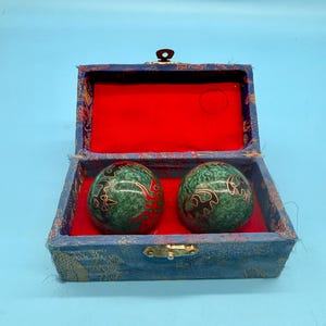 May include: A pair of green and black decorative Chinese health balls inside a blue box with a red interior. The balls have a swirling design with red accents. The box is rectangular with a hinged lid and a clasp closure.