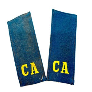 May include: Two vintage blue shoulder boards with yellow embroidered letters "CA". The rectangular boards have a slightly worn appearance, suggesting age. The letters are in a bold, sans-serif font.