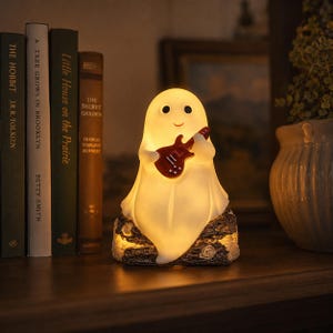 May include: A glowing white ghost figurine with black eyes and a small smile, holding a miniature red electric guitar. The ghost is perched atop a textured, dark brown log-shaped base that emits a warm light from underneath its curved form.