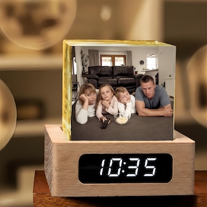 May include: A wooden digital clock with a photo cube displaying a family watching TV. The clock face shows the time 10:35 in white digits. The photo cube sits atop the clock base, showcasing a family portrait.