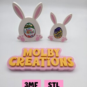 Puede incluir: Dos decoraciones de Pascua con forma de conejito blanco con orejas y patas rosas, cada una con un Kinder Sorpresa y un huevo Cadbury Creme. Debajo, un letrero rosa dice "MOLBY CREATIONS" en letras amarillas. Dos cuadrados rosas dicen "3MF FILE" y "STL FILE".