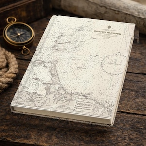 May include: A cream-coloured journal with a map of Boston Harbour printed on the cover. The map is detailed in grey. A brass compass and rope are in the background, suggesting a nautical theme. The journal is on a wooden surface.