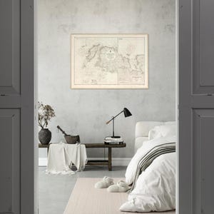 May include: A framed vintage map with a beige border hangs on a gray wall. The map features detailed geographical lines and text. A black desk lamp, a wooden bench, and a bed with white bedding are in the room.