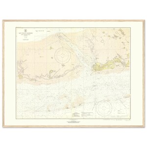 May include: A framed nautical chart of Key West Harbor and Approaches. The chart features detailed lines, symbols, and text, with a color palette of beige, cream, and light yellow. The frame is a light wood color, and the matting is off-white.