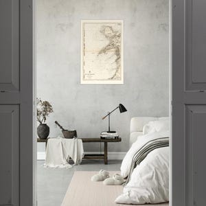 May include: A framed vintage nautical chart in a room setting. The chart is beige with dark brown lines and text, depicting coastal features. The room has a neutral color palette with a bench, lamp, and bed, creating a minimalist aesthetic.