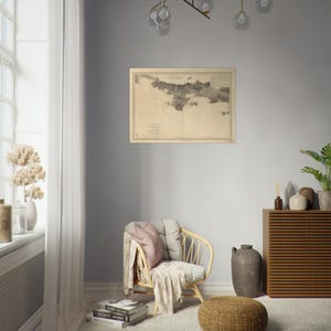 May include: A framed vintage map in neutral tones hangs on a gray wall. The map depicts a coastal region with detailed illustrations. The room features a rattan chair with cushions, a wooden cabinet, and decorative items, creating a cozy atmosphere.