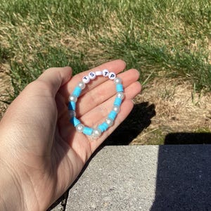 Peut inclure: Un bracelet de perles bleues et blanches, avec les lettres "TS" et "P" épelées en perles de lettres blanches. Le bracelet comporte un charm en forme d'étoile. Le bracelet est tenu dans une main.