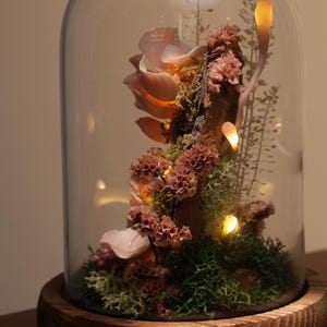 May include: A decorative glass cloche with a wooden base, showcasing a miniature botanical arrangement. The arrangement features pink flowers, green moss, and small lights, creating a whimsical and illuminated display. The cloche is a beautiful home decor item.