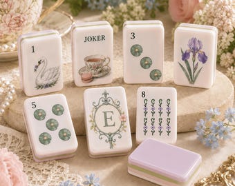 Op de afbeelding: Een set rechthoekige witte tegels met diverse illustraties, waaronder een zwaan, een theekopje, waterlelies, een iris en de letter E. Eén tegel is gelabeld als JOKER, terwijl andere de cijfers 1, 3, 5 en 8 tonen. Een effen lavendelkleurige tegel.