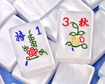 May include: Two white rectangular mahjong tiles with a pearlescent finish. The left tile features a blue number 1, a red flower, and green stems. The right tile displays a red number 3, the red kanji for autumn, and a green and yellow plant design.