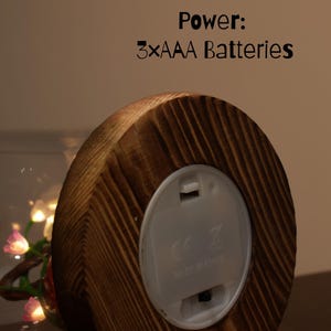 May include: A wooden base with a round, light-colored plastic battery compartment. The text "Power: 3xAAA Batteries" is visible. The wood has a natural grain pattern. A glass sphere with fairy lights is partially visible.
