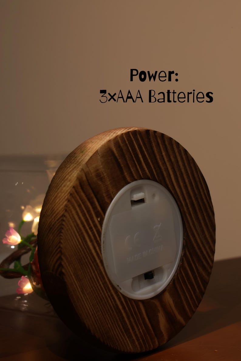 May include: A wooden base with a round, light-colored plastic battery compartment. The text "Power: 3xAAA Batteries" is visible. The wood has a natural grain pattern. A glass sphere with fairy lights is partially visible.