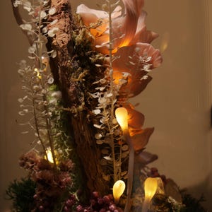 May include: A decorative glass cloche featuring a miniature illuminated woodland scene. The scene includes a piece of wood, moss, small pink berries, and glowing mushroom-shaped lights. The background is a warm, neutral tone.