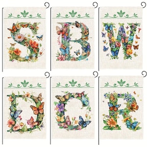 May include: Garden flags featuring letters S, B, W, G, C, and R, each adorned with colorful butterflies and flowers. The flags have a beige background with a green leaf design at the top and are designed for outdoor use.