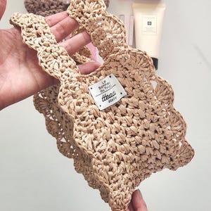 May include: A small, beige woven tote bag with scalloped edges and handles. The bag features a rectangular tag with the text "Le Bonheur du Tricot" and "thead atelier". The bag is made of a natural fibre material.