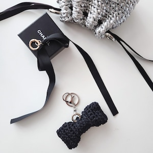 May include: A black crocheted bow keychain with a silver clasp. A small black box with gold accents and a black ribbon are also present. A silver woven bag with a drawstring closure is in the background.