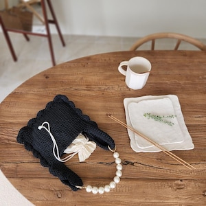 May include: A navy blue crocheted tote bag with a scalloped edge and a pearl handle. The bag is open, revealing a white drawstring pouch inside. A white mug and two white square plates with a sprig of greenery and chopsticks are on a wooden table.