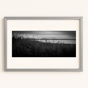 Könnte beinhalten: Schwarz-Weiß-Landschaftsfotografie in einem silbernen Rahmen. Das Bild zeigt hohes Gras im Vordergrund, ein Gewässer und einen bewölkten Himmel. Das Foto ist mit einem weißen Passepartout gerahmt.