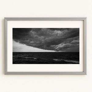 May include: A framed black and white photograph of a stormy seascape. Dark, ominous clouds dominate the sky above the turbulent ocean. The photograph is encased in a silver-colored frame, adding a modern touch to the dramatic scene.