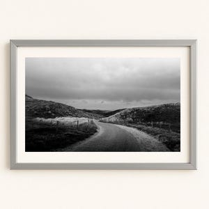 Könnte beinhalten: Schwarz-Weiß-Fotografie in einem Rahmen, die eine kurvenreiche Straße durch eine hügelige Landschaft unter einem bewölkten Himmel zeigt. Die Straße schlängelt sich in die Ferne, flankiert von grasbewachsenen Hügeln und einem Drahtzaun. Der Rahmen ist silberfarben.
