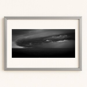 Puede incluir: Fotografía en blanco y negro enmarcada de una tormenta dramática sobre el océano. La imagen presenta una gran formación de nubes oscuras que dominan el cielo, con el mar debajo. La fotografía está encerrada en un marco plateado.