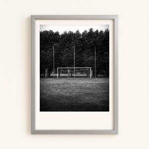 Peut inclure: Photographie encadrée en noir et blanc d'un terrain de football. L'image présente un but au centre, avec des arbres en arrière-plan. La photographie est dans un cadre argenté et est affichée sur un mur blanc.