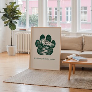 May include: A framed print with a dark green paw print design, depicting a mountain and forest scene. The print includes the text "It's not the path-it's the partner". The artwork is displayed in a bright room with a neutral color scheme.