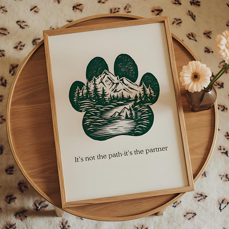 May include: Framed print featuring a green paw print design with a mountain and forest landscape. The print includes the text "It's not the path-it's the partner" in a wooden frame. The artwork is displayed on a round wooden table.
