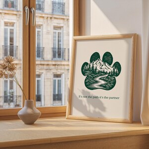 May include: A framed print with a dark green paw print design, depicting a mountain and forest scene. The text "It's not the path-it's the partner" is printed below. The print is displayed on a wooden surface, near a window.